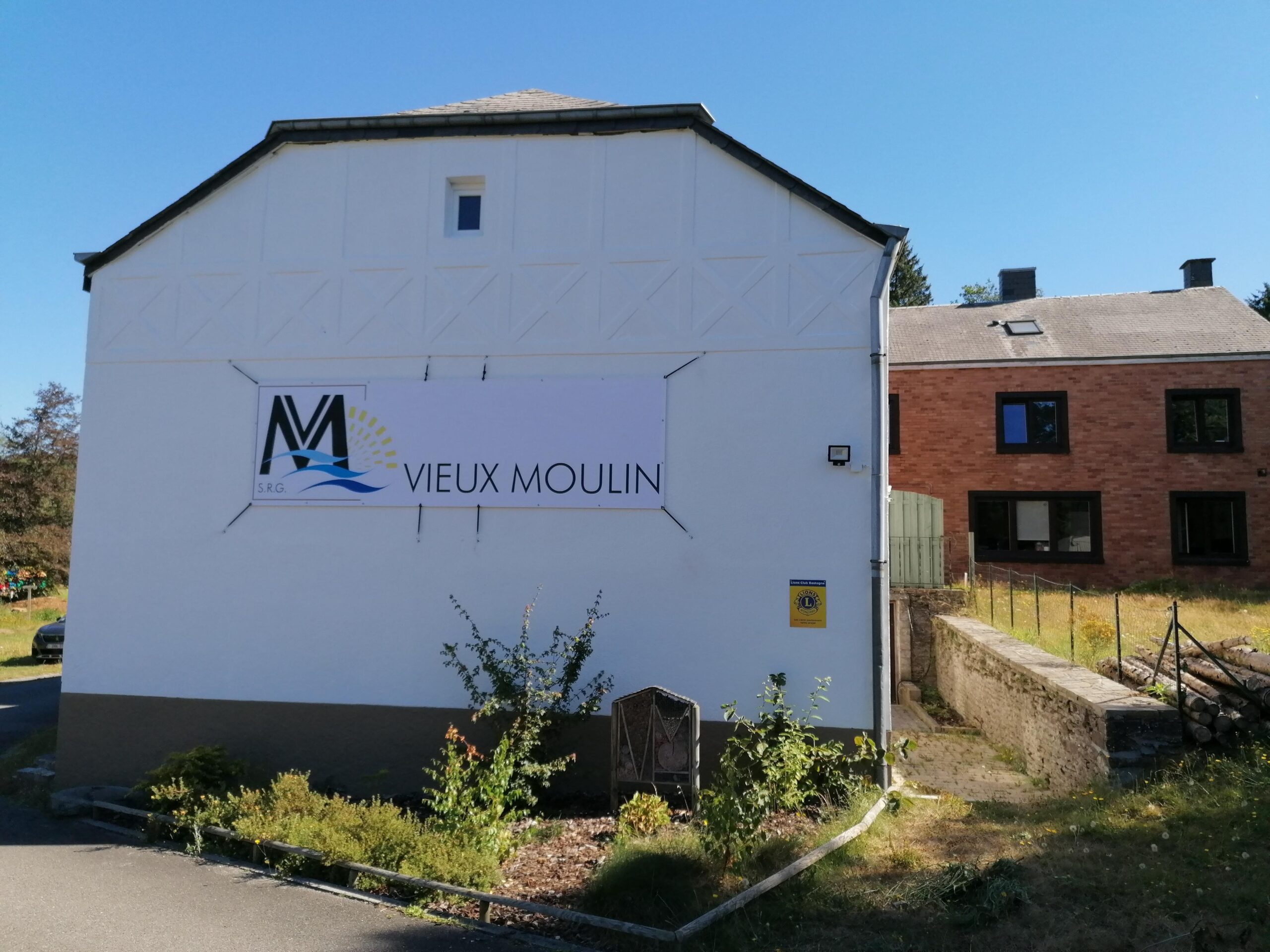 Devanture de la maison le vieux moulin avec une banderole accrochée à celle-ci contenant le nom de la maison et le logo de l'ASBL